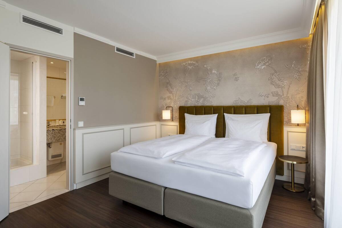 Une chambre d’hôtel avec un lit double, une petite table de chevet, une lampe fixée au mur et une salle de bain visible à travers une porte ouverte.