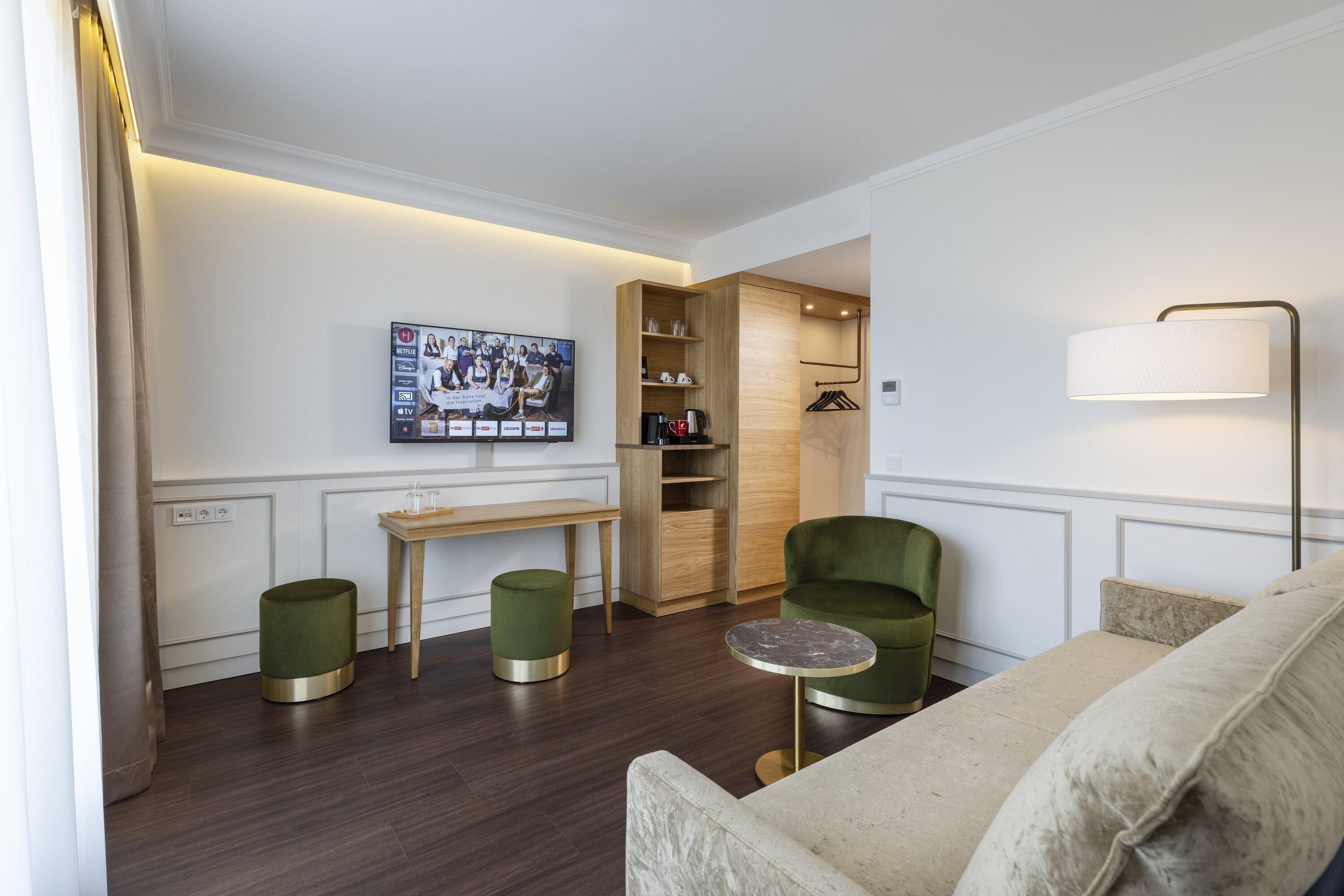 A modern hotel room with a green armchair, a beige sofa, a wooden cabinet, and a television mounted on the wall.