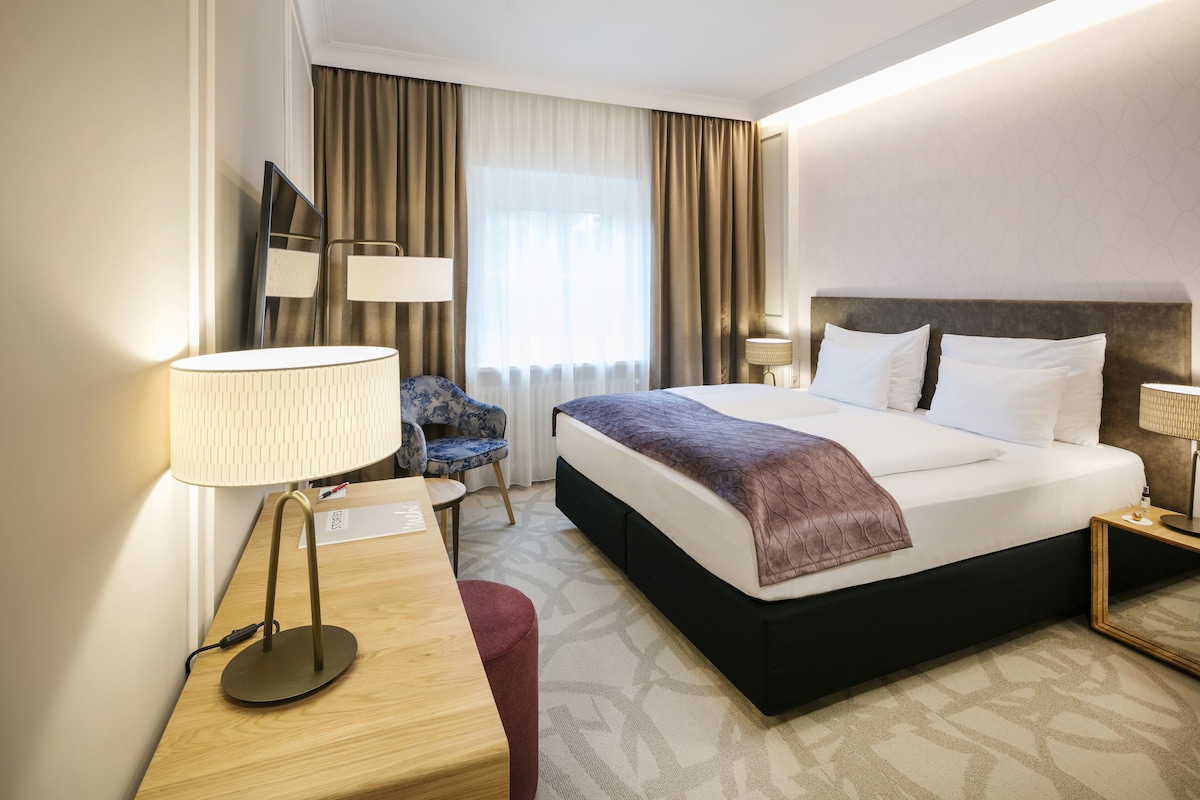 Ein Hotelzimmer mit Bett, Nachttischen, Schreibtisch, Stuhl, Lampe und einem fenster mit Vorhängen.