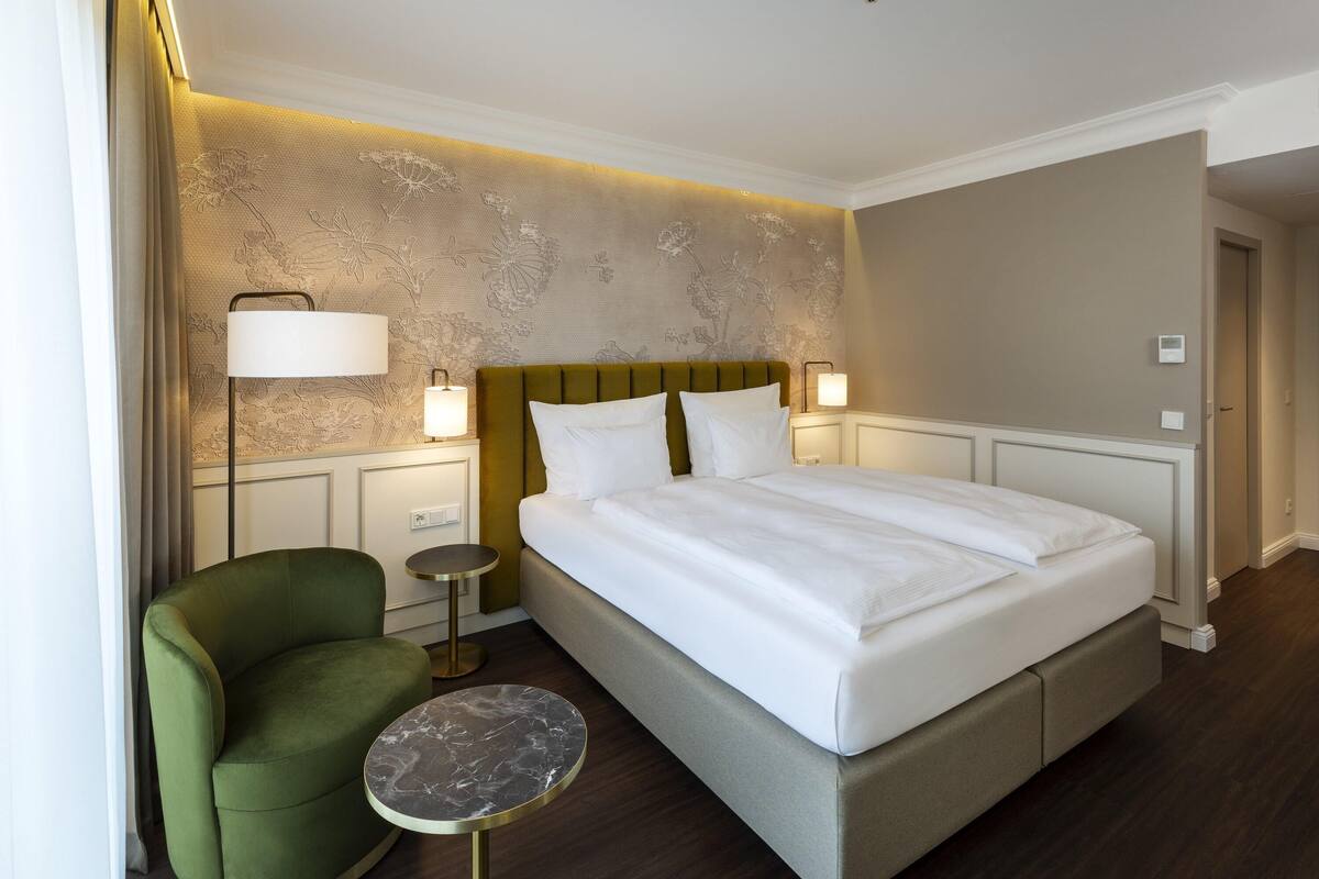 Une chambre d’hôtel avec un grand lit, un fauteuil vert, une petite table ronde et un mur recouvert d’un papier peint à motifs floraux.