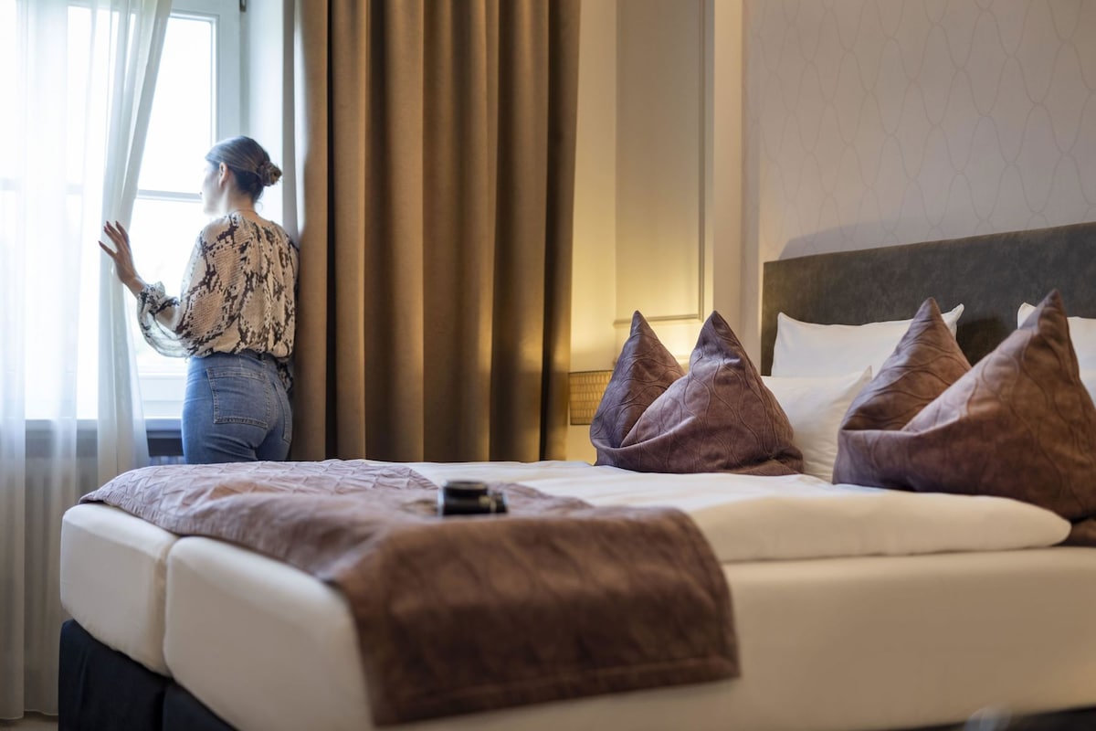 Ein Hotelzimmer mit Bett, Kissen, einem Nachttisch und einem Fenster mit Vorhängen.