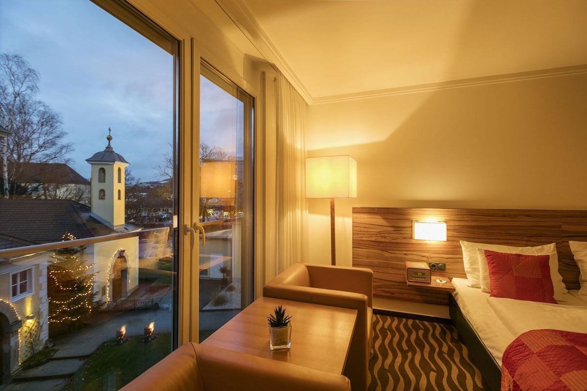 Ein Hotelzimmer mit Bett, Schreibtisch, Stuhl, Lampe und Blick auf eine Kirche sowie Weihnachtsbeleuchtung vor den Fenstern.