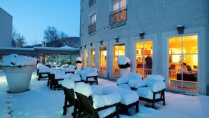 Un bâtiment doté de nombreuses fenêtres et d’un espace extérieur aménagé avec des sièges, le tout recouvert de neige.