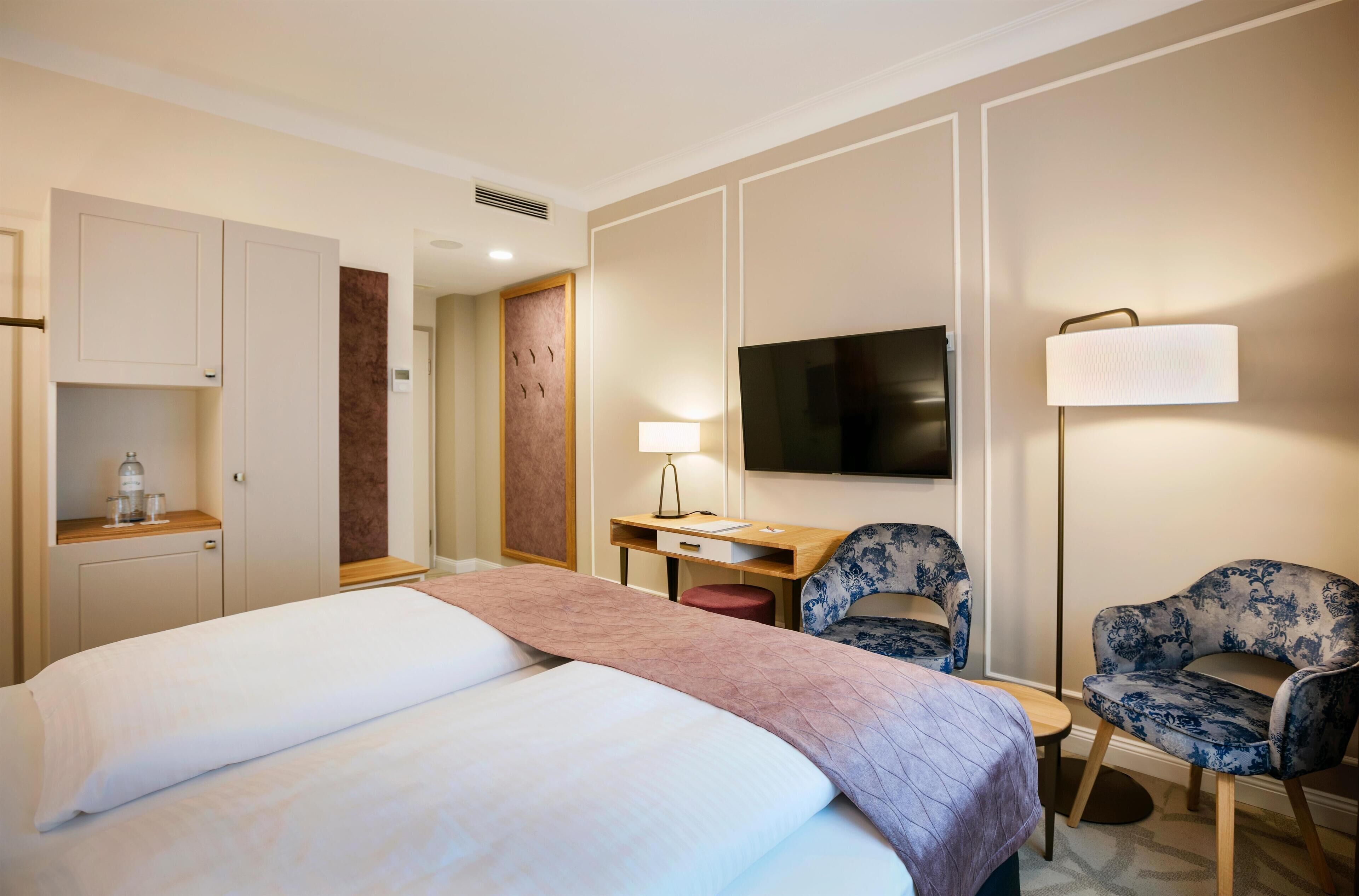 A hotel room with a bed, a desk, a chair, a TV, and a lamp.