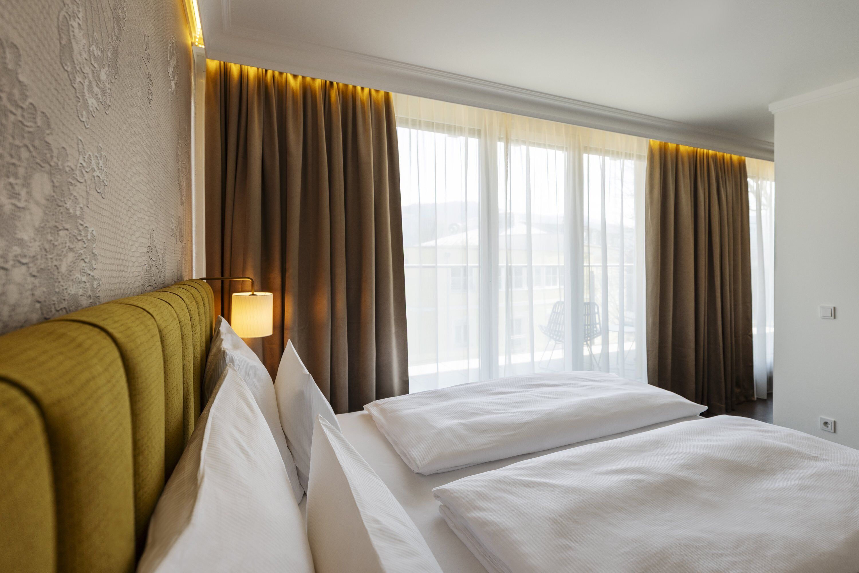 Una habitación de hotel con una cama grande, cabecera, lámpara de noche y una ventana con cortinas.