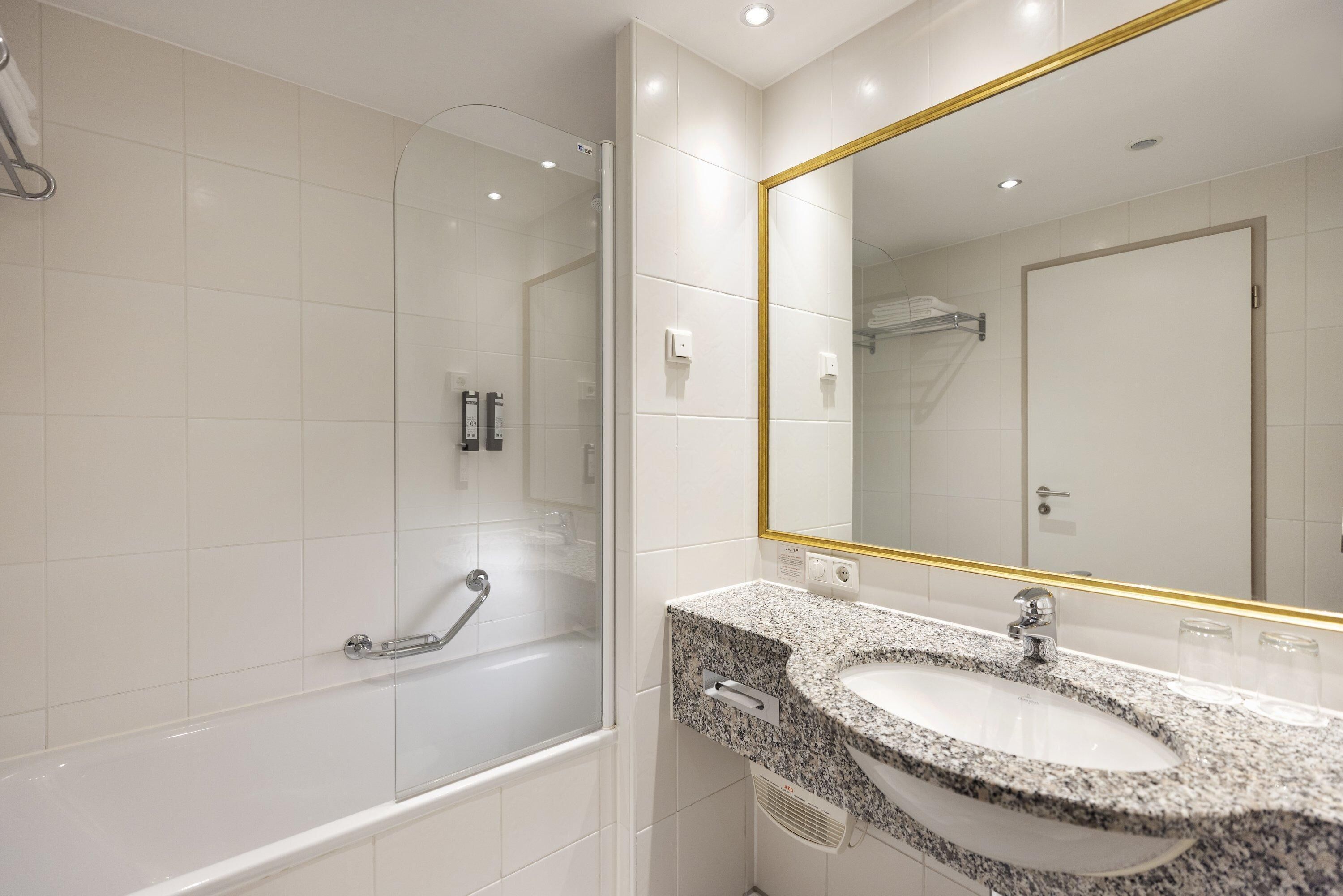 Un baño moderno con ducha envuelta en vidrio, lavamanos de granito y un espejo grande con marco dorado.