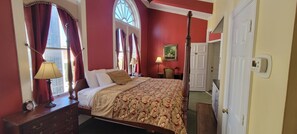 Classic four-poster bed with a patterned bedspread and two pillows, a nightstand with a lamp and alarm clock, and a large window with a painting on the wall.