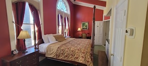 Classic four-poster bed with a patterned bedspread and two pillows, a nightstand with a lamp and alarm clock, and a large window with a painting on the wall.