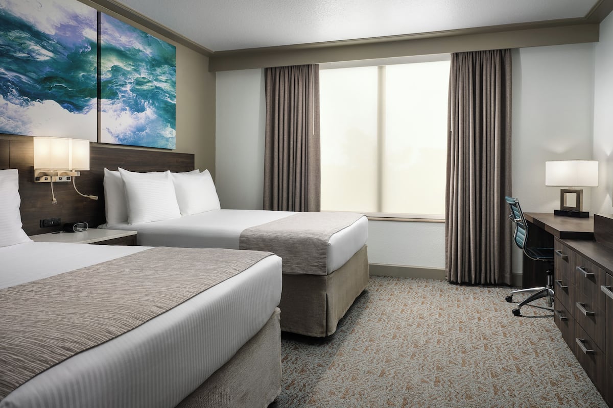 A hotel room with two beds, a desk, a chair, a lamp, and a large window with curtains.