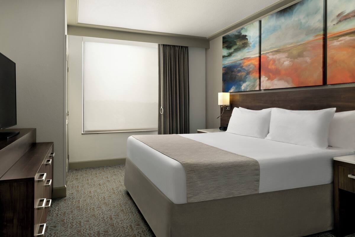 A hotel room with a large bed, a TV, a desk, and a painting on the wall.
