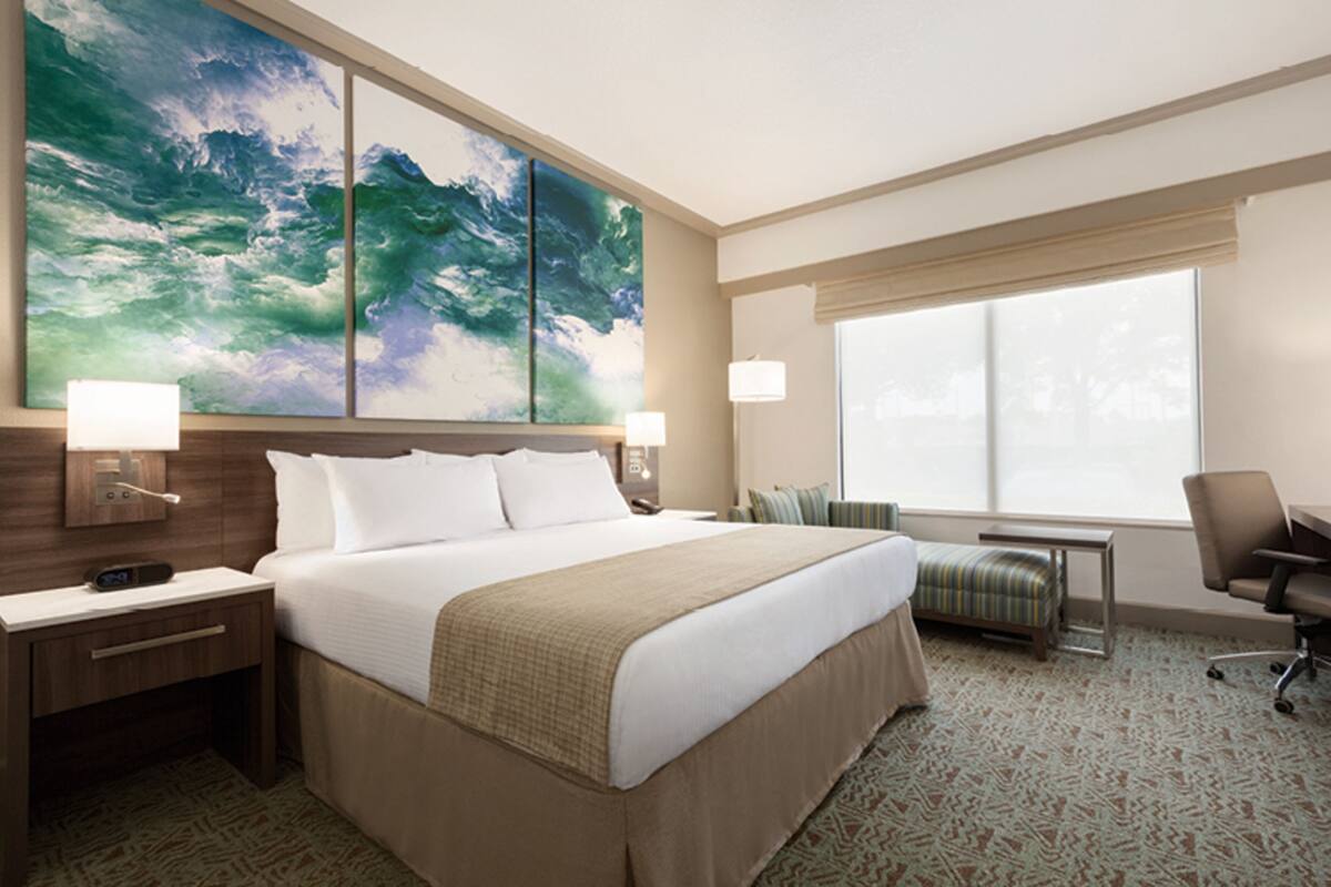 A hotel room with a large bed, two bedside tables, a desk, a chair, a window, and a large abstract painting on the wall.