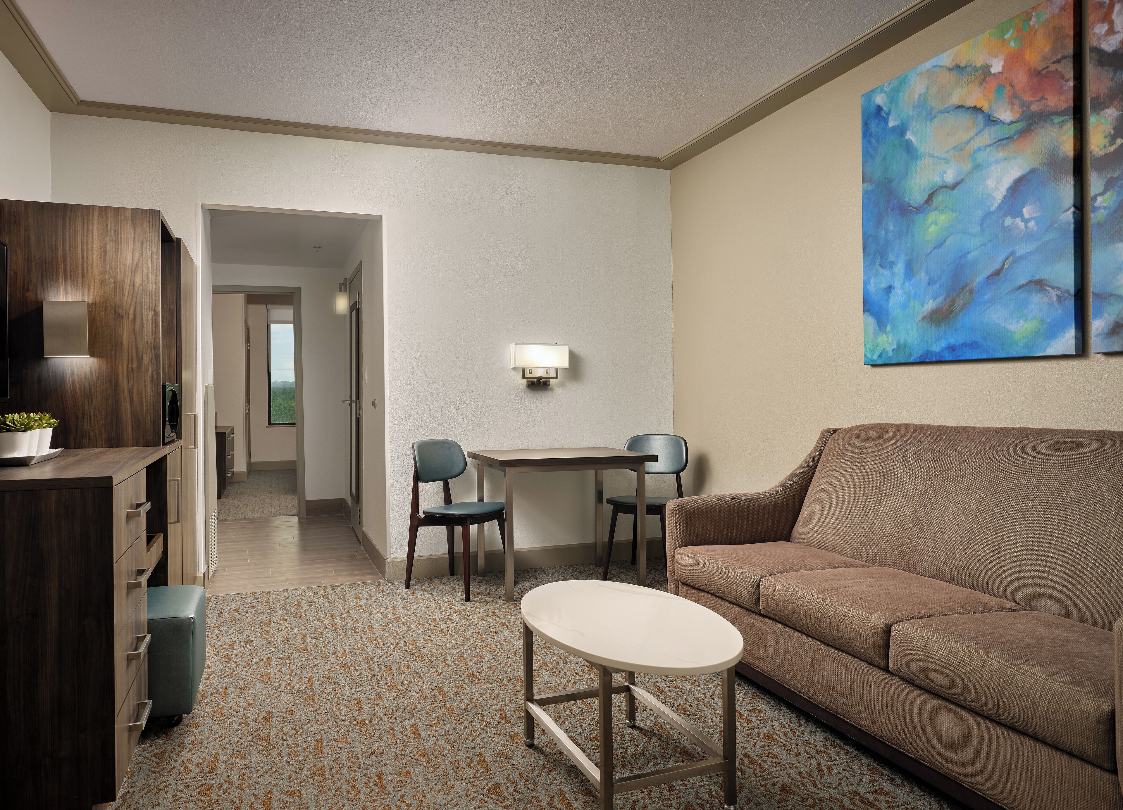 Una habitación de hotel moderna con un sofá marrón, una mesita redonda pequeña, dos sillas, un escritorio y un cuadro abstracto grande en la pared.