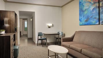 A modern hotel room with a brown sofa, a small round table, two chairs, a desk, and a large abstract painting on the wall.