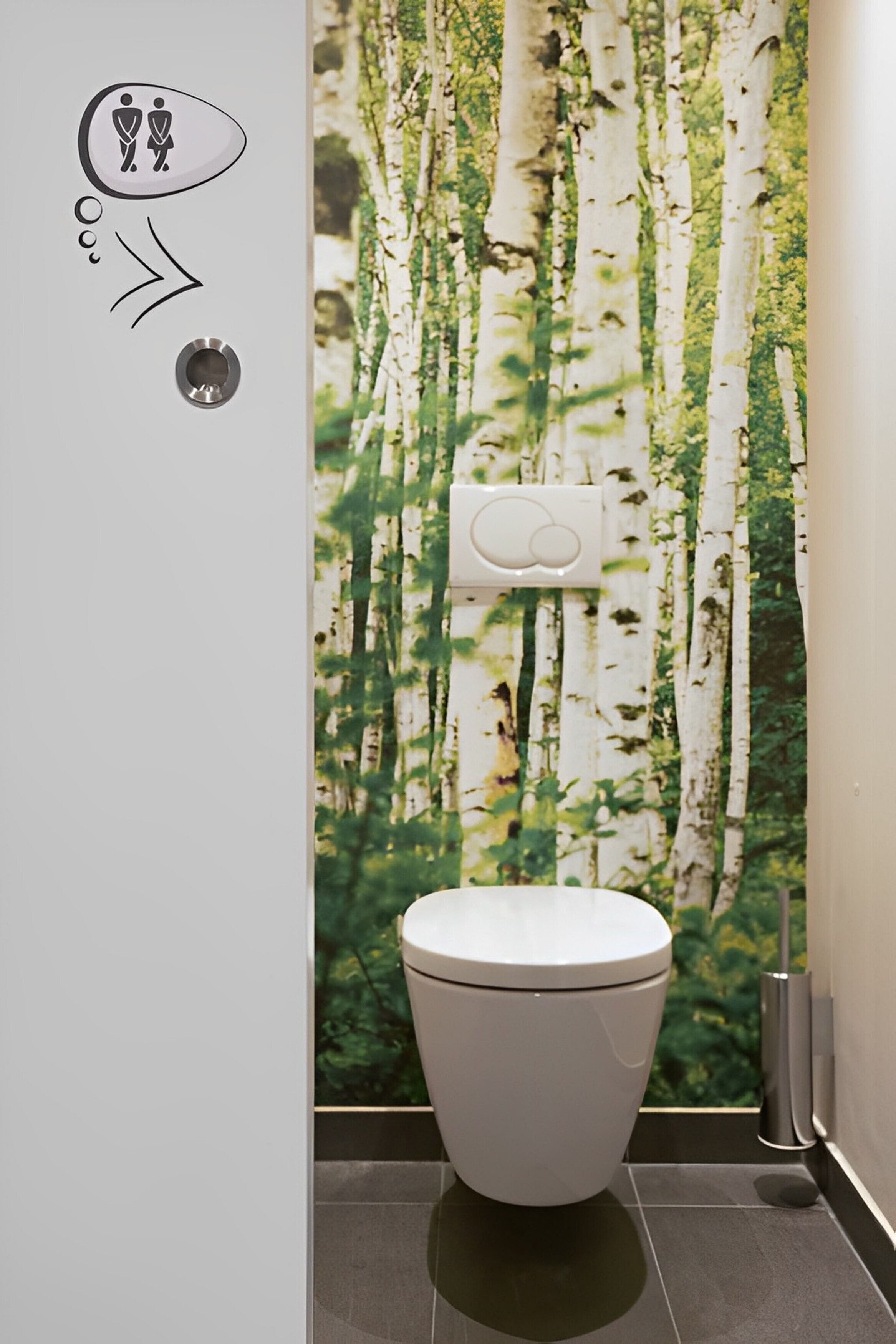 A toilet with a white lid, a wall-mounted flush button, and a wall decal of birch trees.