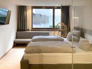 A modern hotel room with a large bed, a seating area, a television, and a window with curtains.