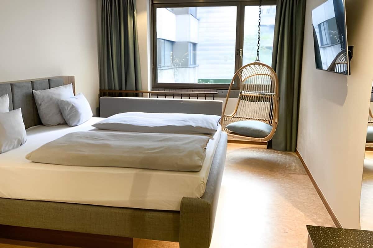 A hotel room with a bed, a hanging chair, a TV mounted on the wall, and a window with curtains.