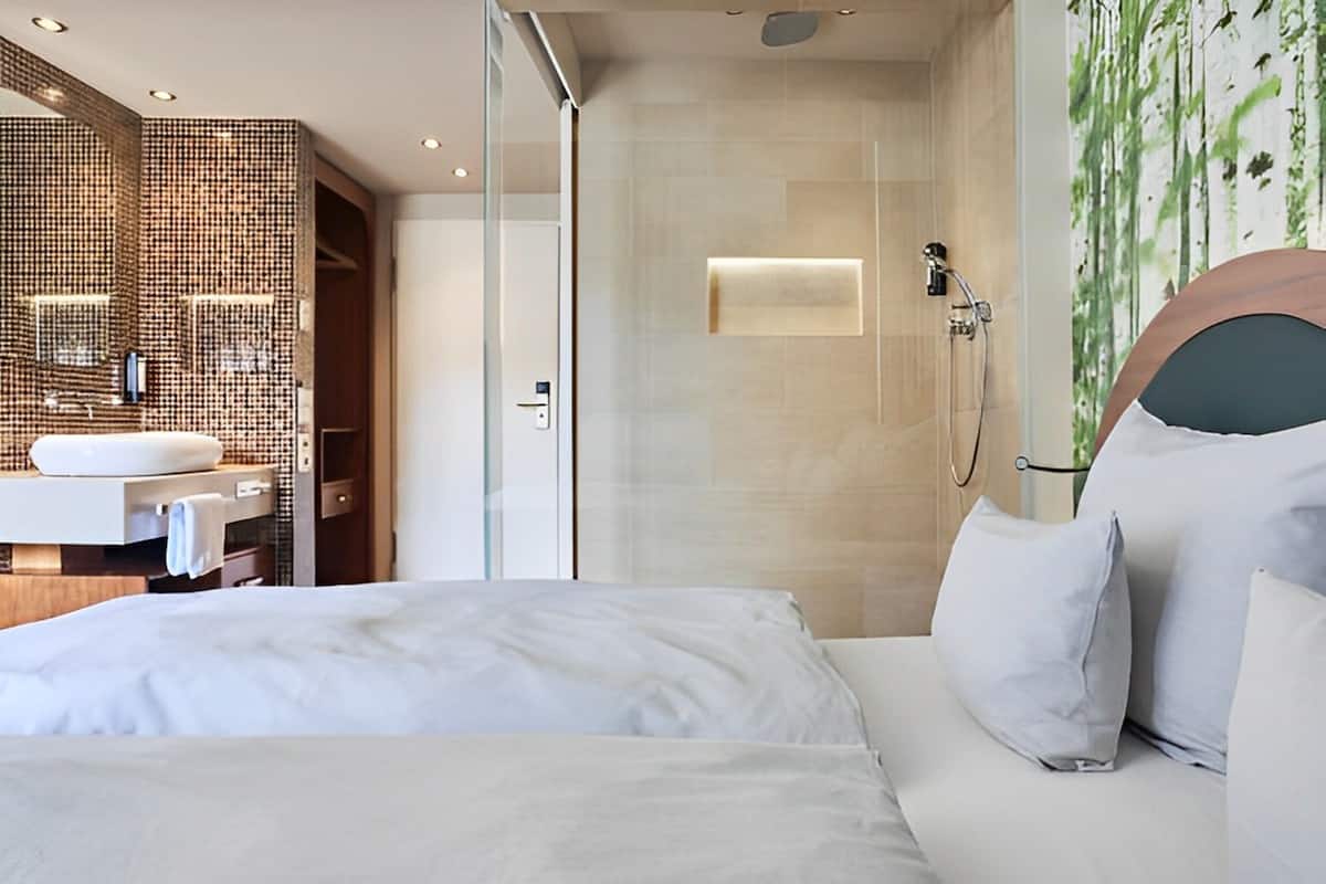 A modern hotel room with a large bed, a bathroom with a glass shower enclosure, and a wall-mounted sink.