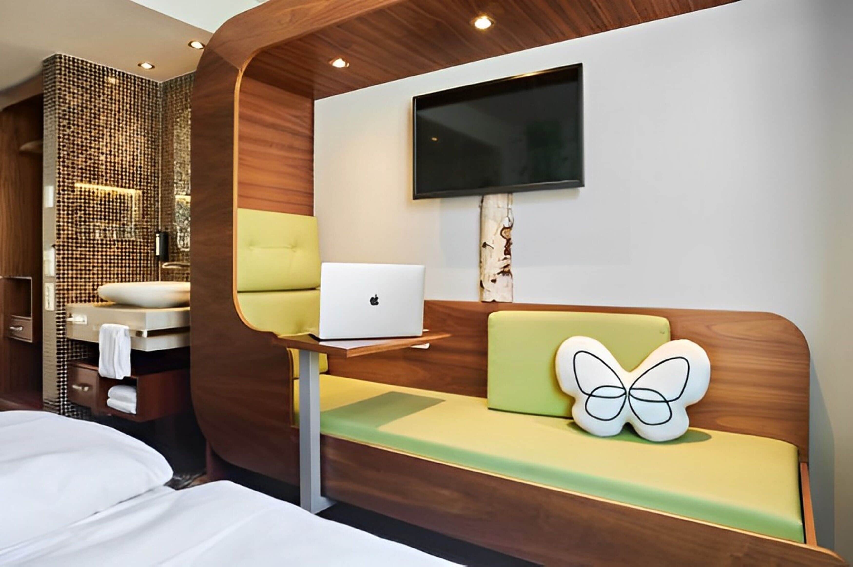 A modern hotel room with a wooden accent wall, a flat-screen TV mounted above a green sofa, and a laptop on a small table.
