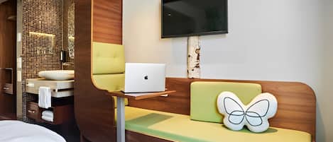 Una habitación de hotel moderna con una pared decorada con madera, un televisor de pantalla plana sobre un sofá verde y un ordenador portátil sobre una mesita.