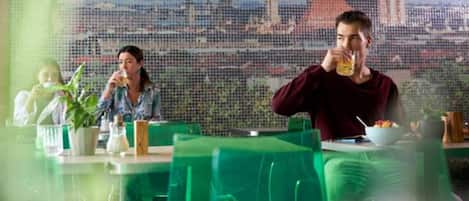 A dining area with green chairs and tables, a cityscape view through a patterned window, and a person drinking from a glass.