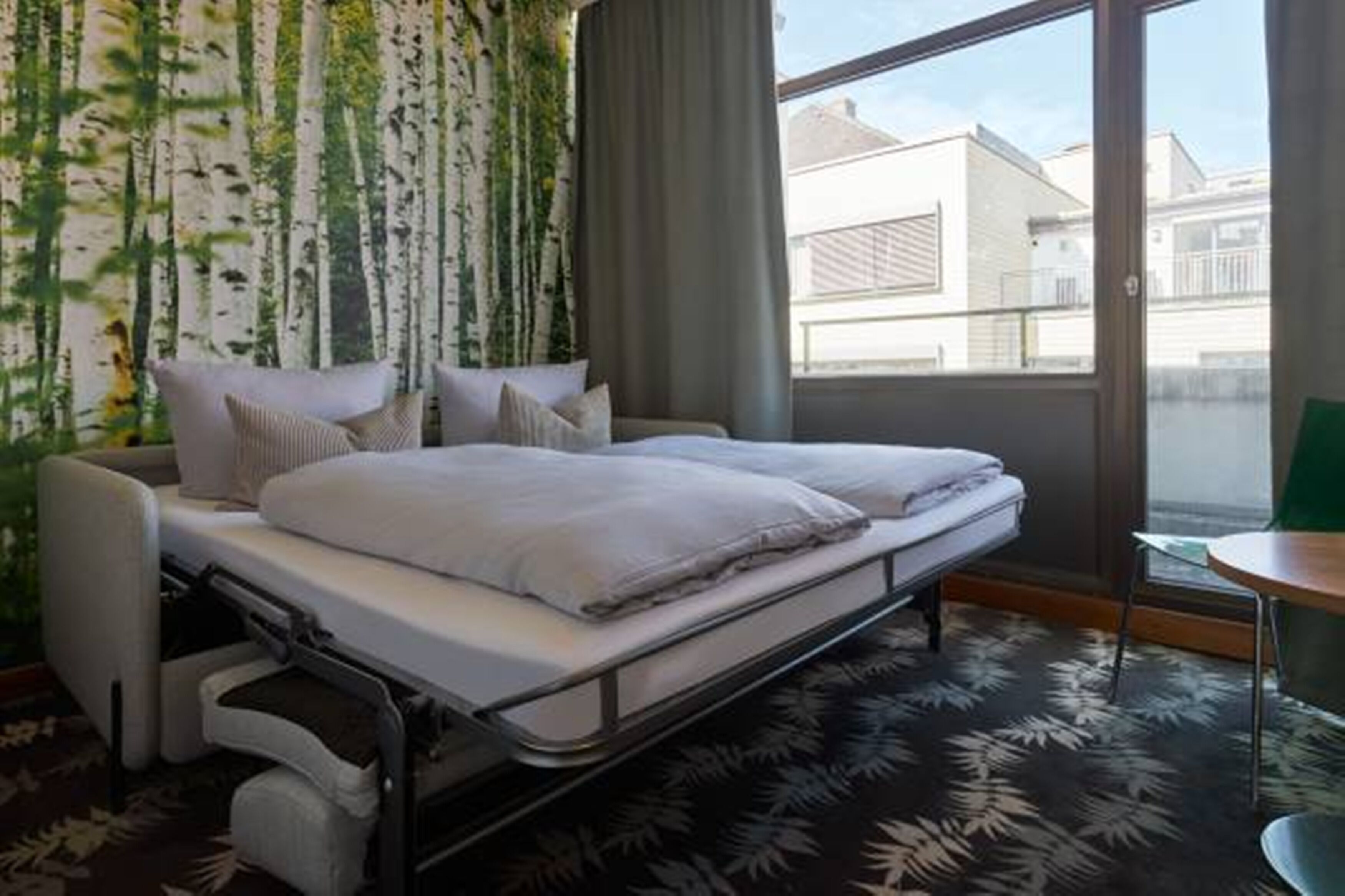 A hotel room with a bed, a chair, a table, and a view of buildings through a window.