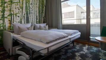 A hotel room with a bed, a chair, a table, and a view of buildings through a window.