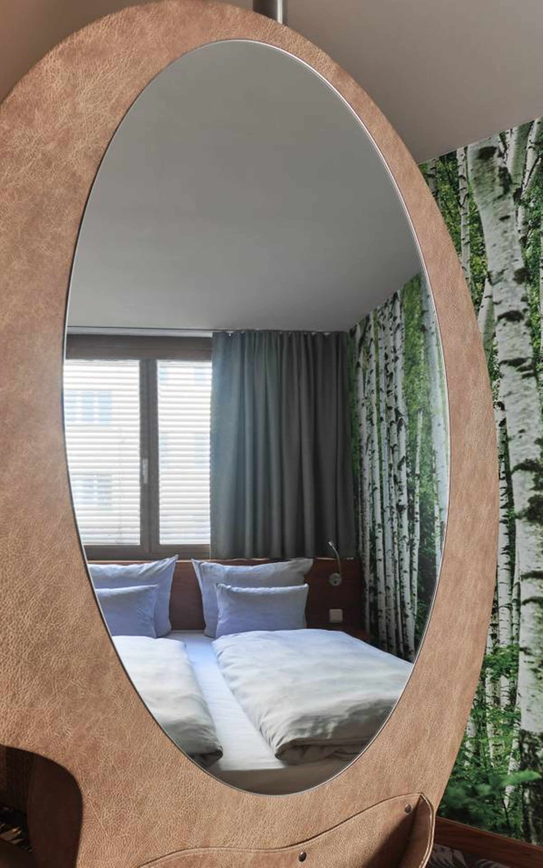Una habitación de hotel con un espejo ovalado grande, una cama con ropa blanca, una ventana con persianas y una cortina con estampado de árboles de abedul.