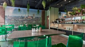 Un restaurante con sillas y mesas verdes, un mural de paisaje urbano en la pared y plantas colgantes.