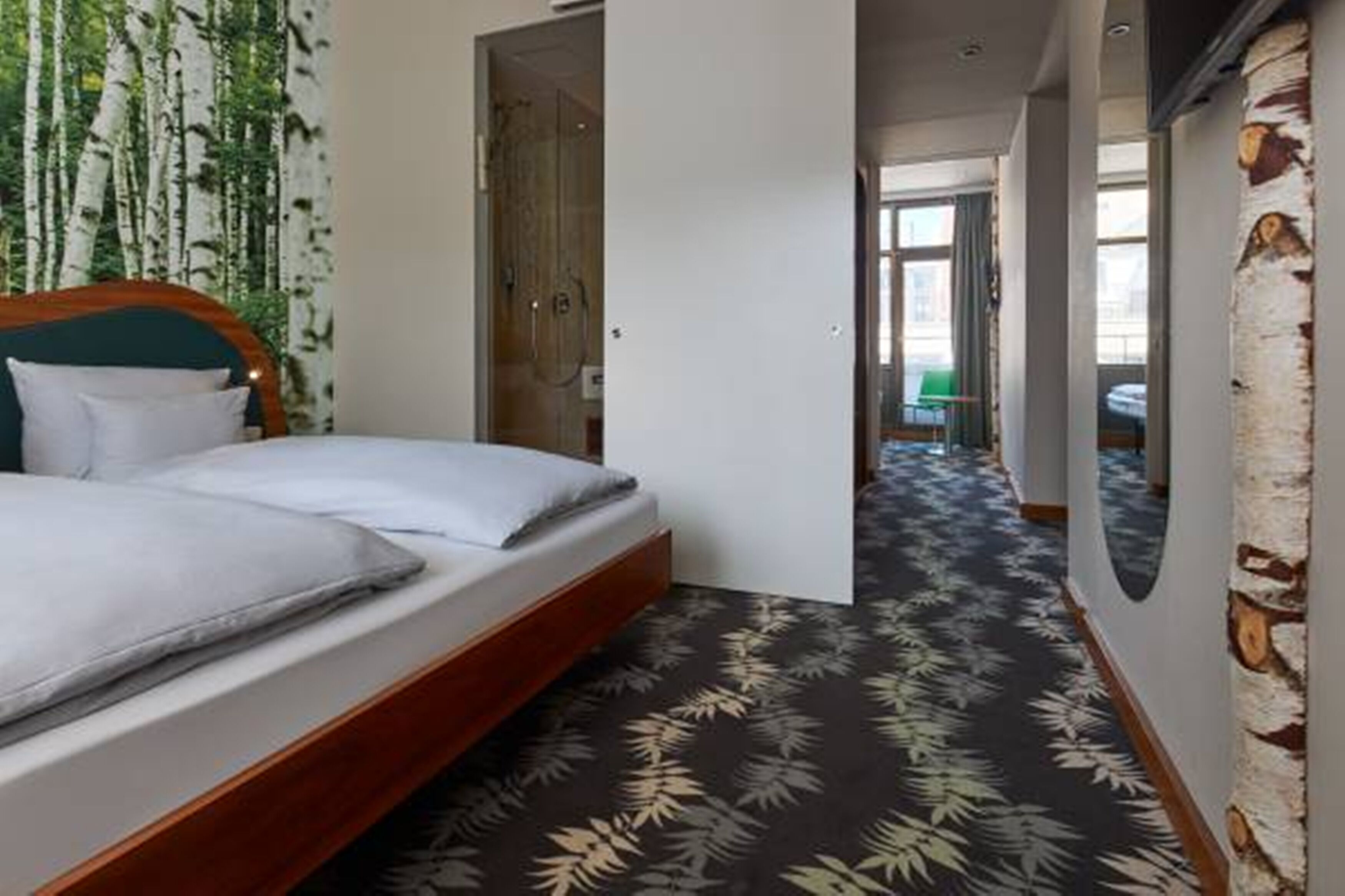 A hotel room with a bed, a door leading to another room, and a hallway with a patterned carpet.