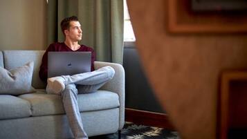 Una persona sentada en un sofá gris con una computadora portátil, en una habitación con un tapete estampado y una ventana con cortinas.