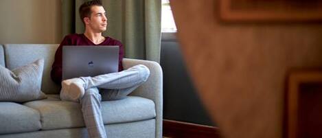 A person sitting on a grey sofa with a laptop, in a room with a patterned rug and a window with curtains.