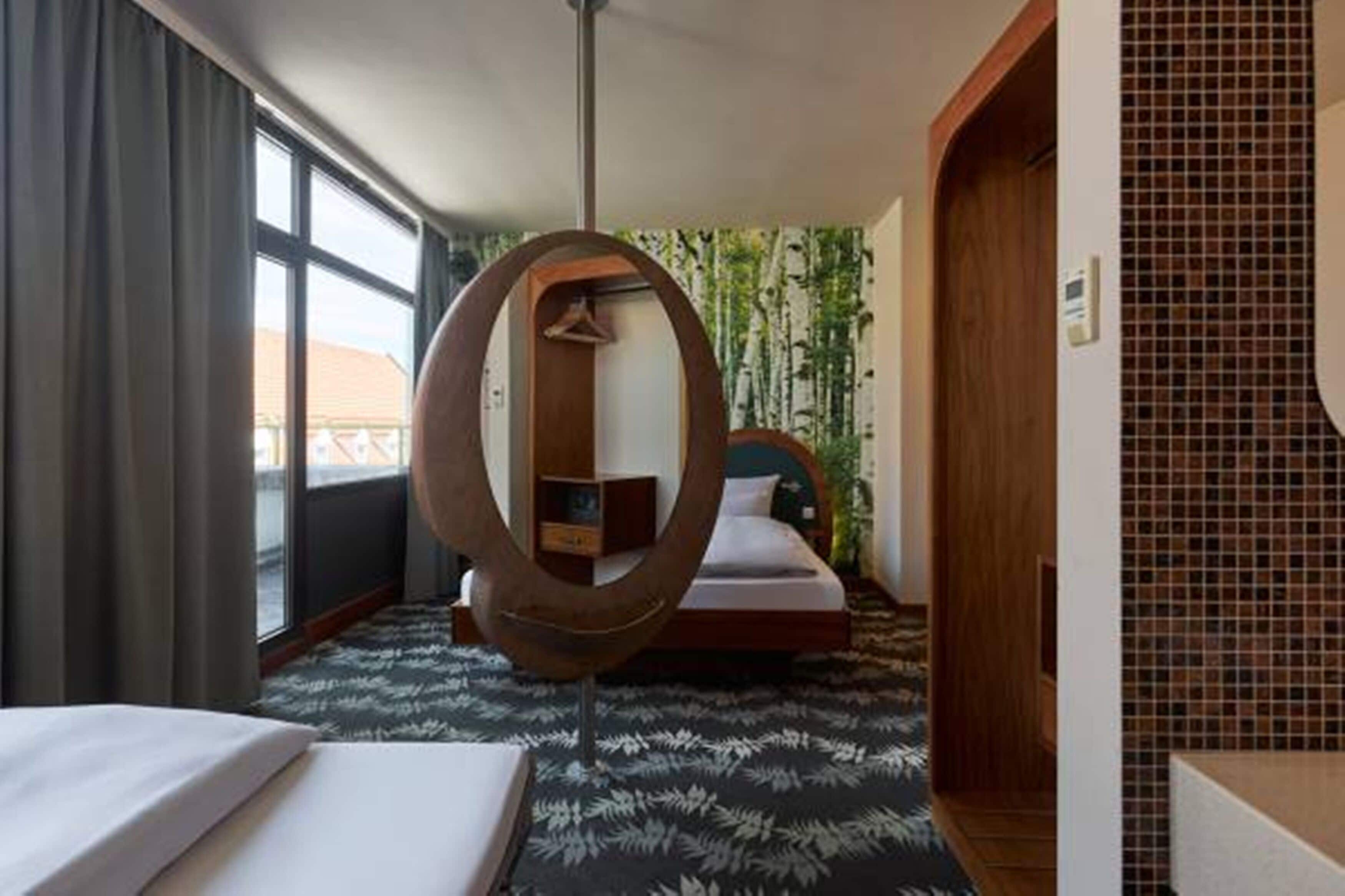 A hotel room with a bed, a circular mirror, a wardrobe, and a view of the outside through a window.