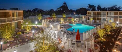 Zona de piscina exterior bien iluminada con sillones de descanso y sombrillas, rodeada de jardines ajardinados y edificios modernos.