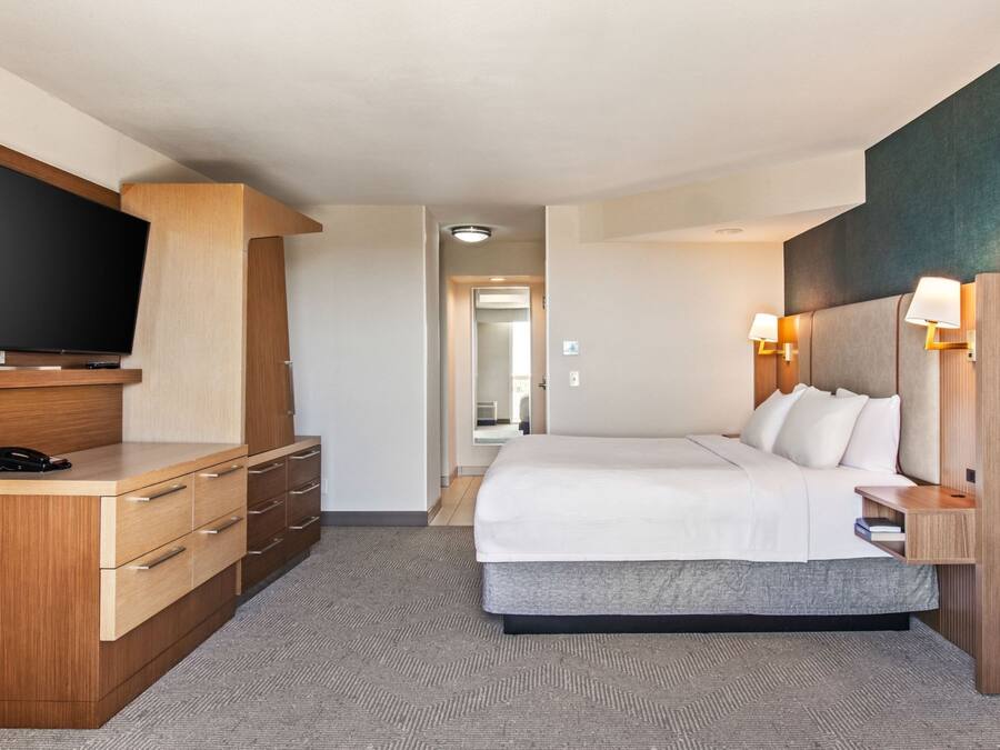 A hotel room with a bed, a TV mounted on a wooden dresser, a desk with drawers, a bedside table with a lamp, and a carpeted floor.