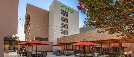 An outdoor patio area with tables and chairs, string lights, and a building with a sign that reads "Holiday Inn Express."