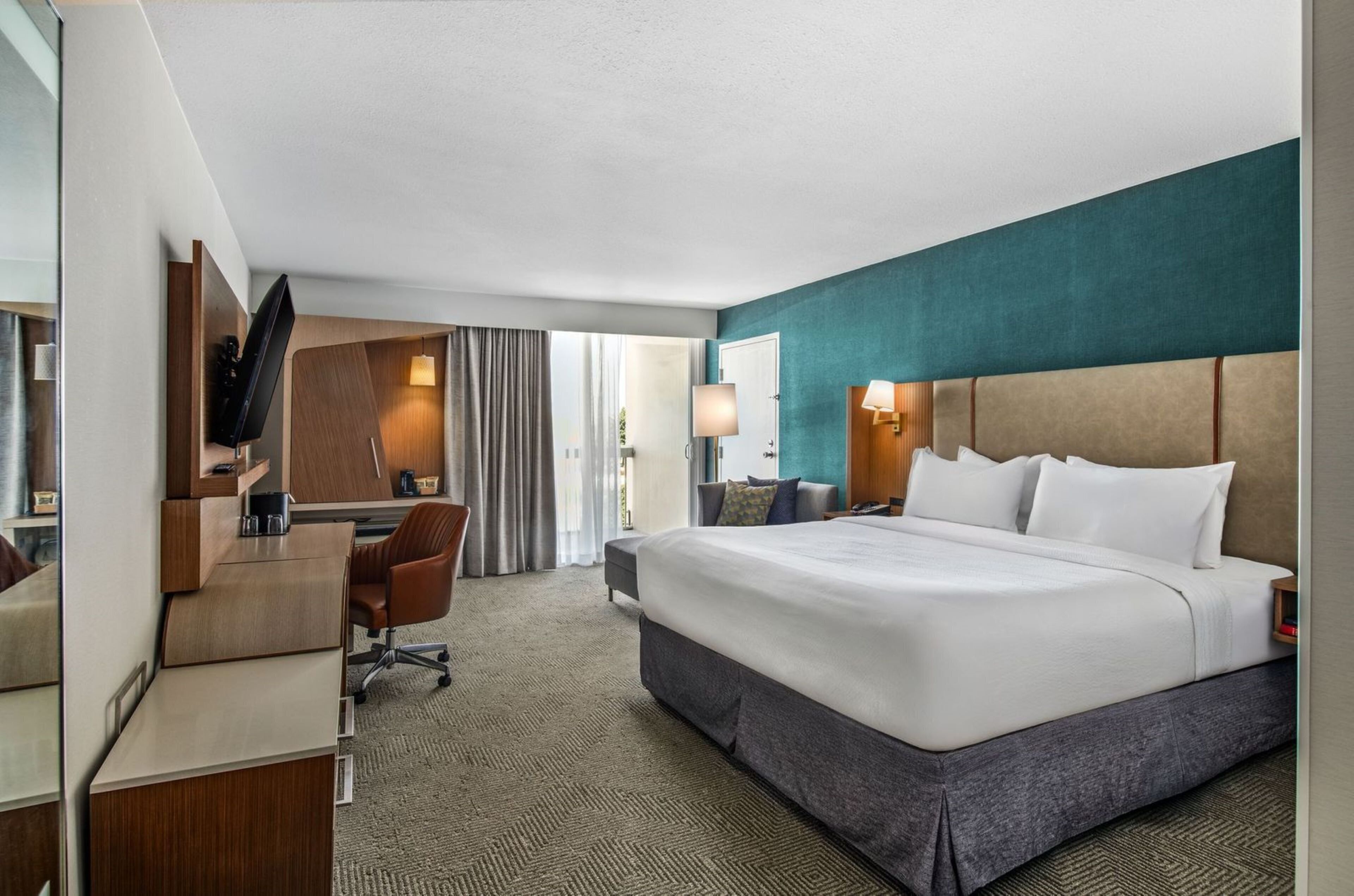 A hotel room with a large bed, a desk, a chair, a TV, and a window with curtains.