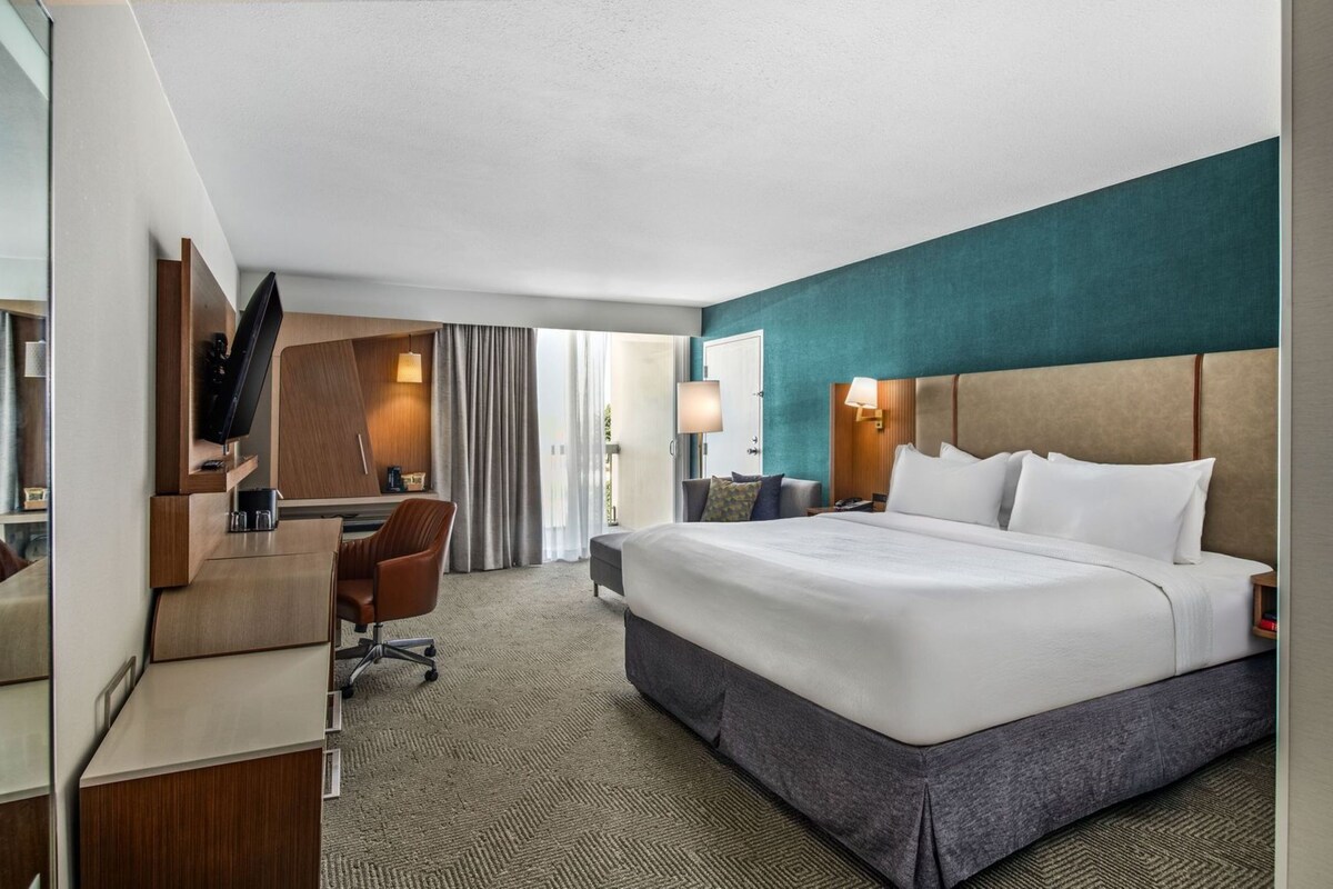 A hotel room with a large bed, a desk, a chair, a TV, and a window with curtains.