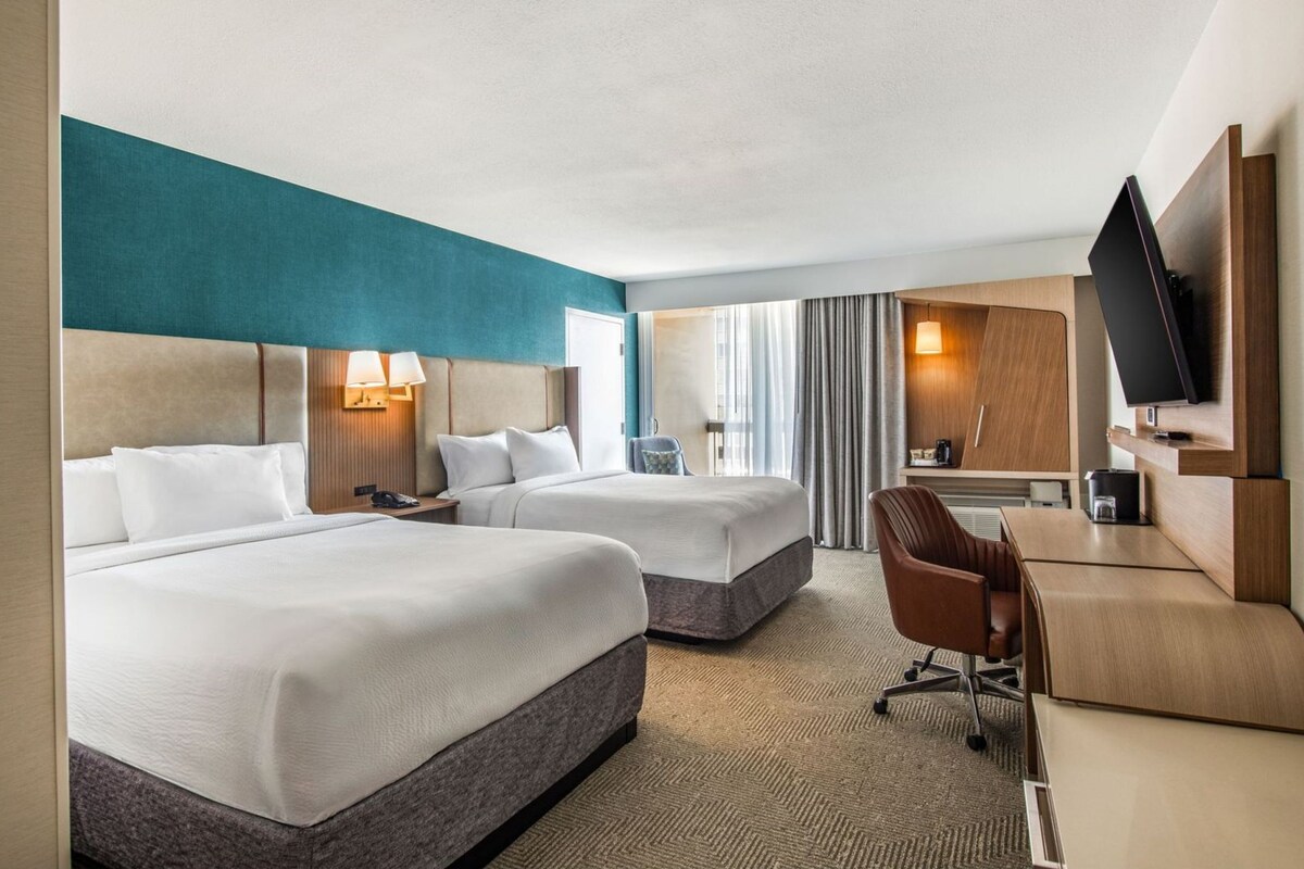 A hotel room with two beds, a desk, a chair, a TV, and a large window with curtains.