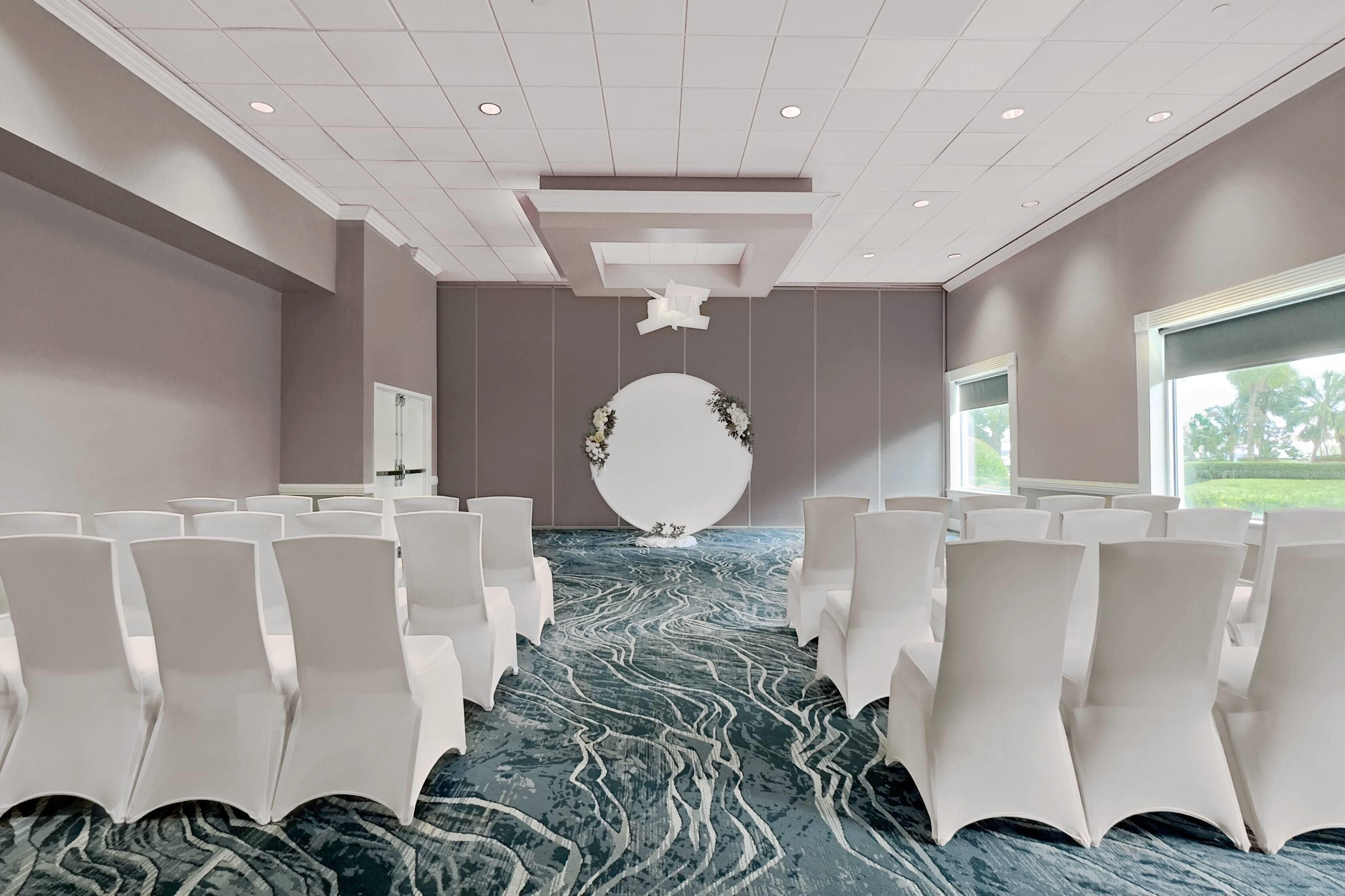 Un salón de conferencias con filas de sillas blancas, una alfombra con diseño de mármol y una gran pieza decorativa blanca en la pared.