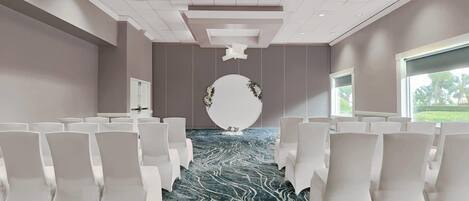 Un salón de conferencias con filas de sillas blancas, una alfombra con diseño de mármol y una gran pieza decorativa blanca en la pared.