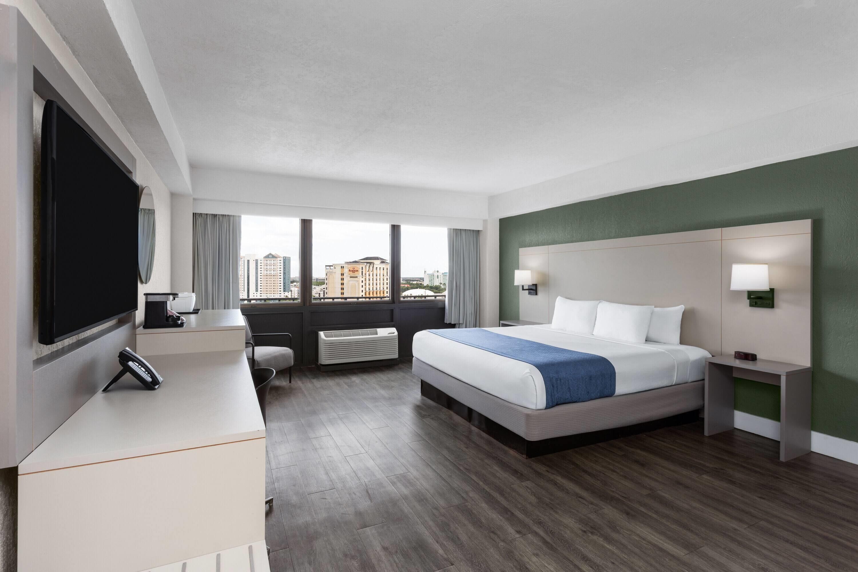 A hotel room with a bed, a desk, a TV, a window with a city view, and a wall-mounted lamp.