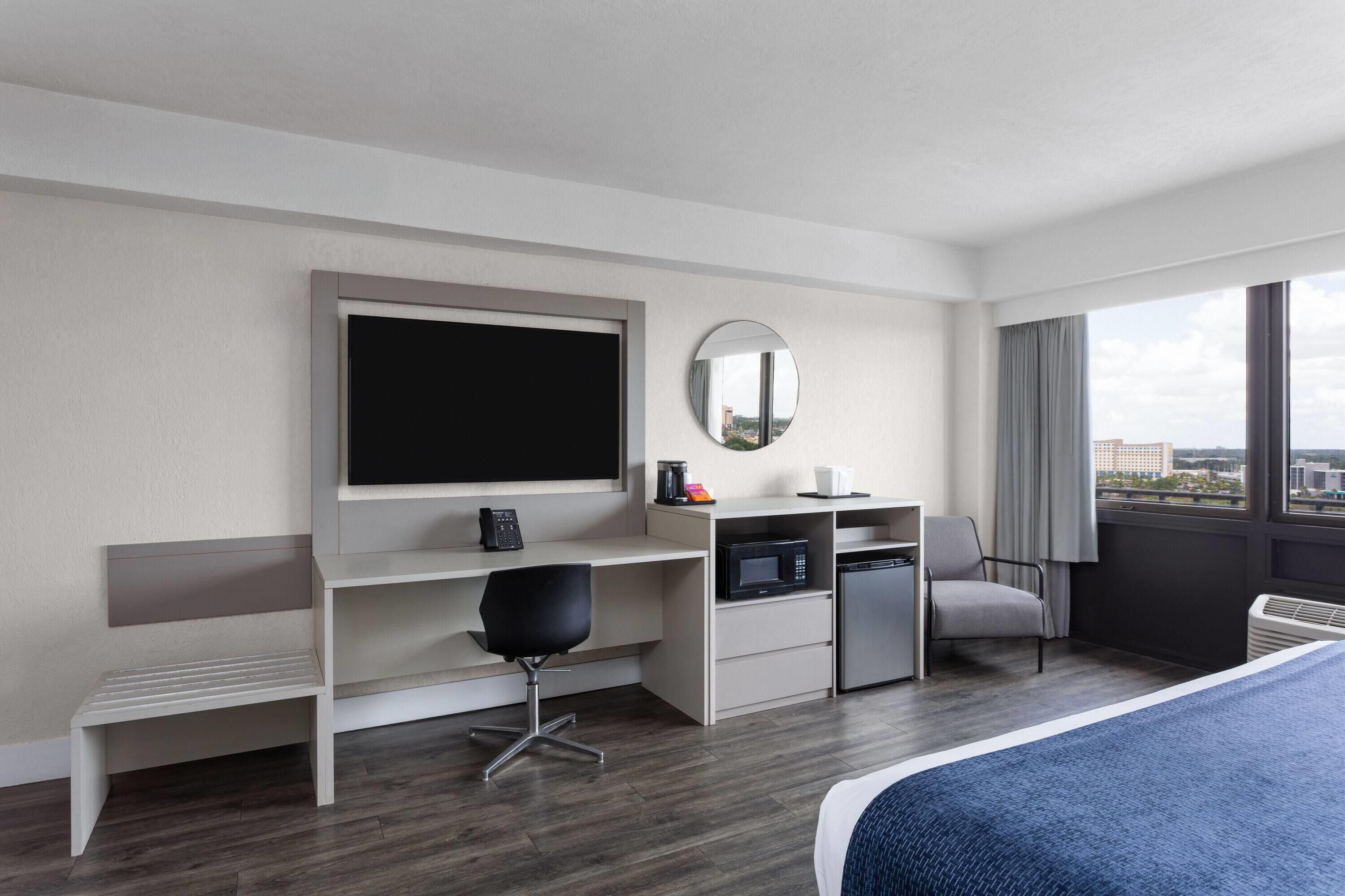 Une chambre d’hôtel comprenant un lit, un bureau avec une chaise, une télévision, un four à micro-ondes et une grande fenêtre donnant sur la ville.