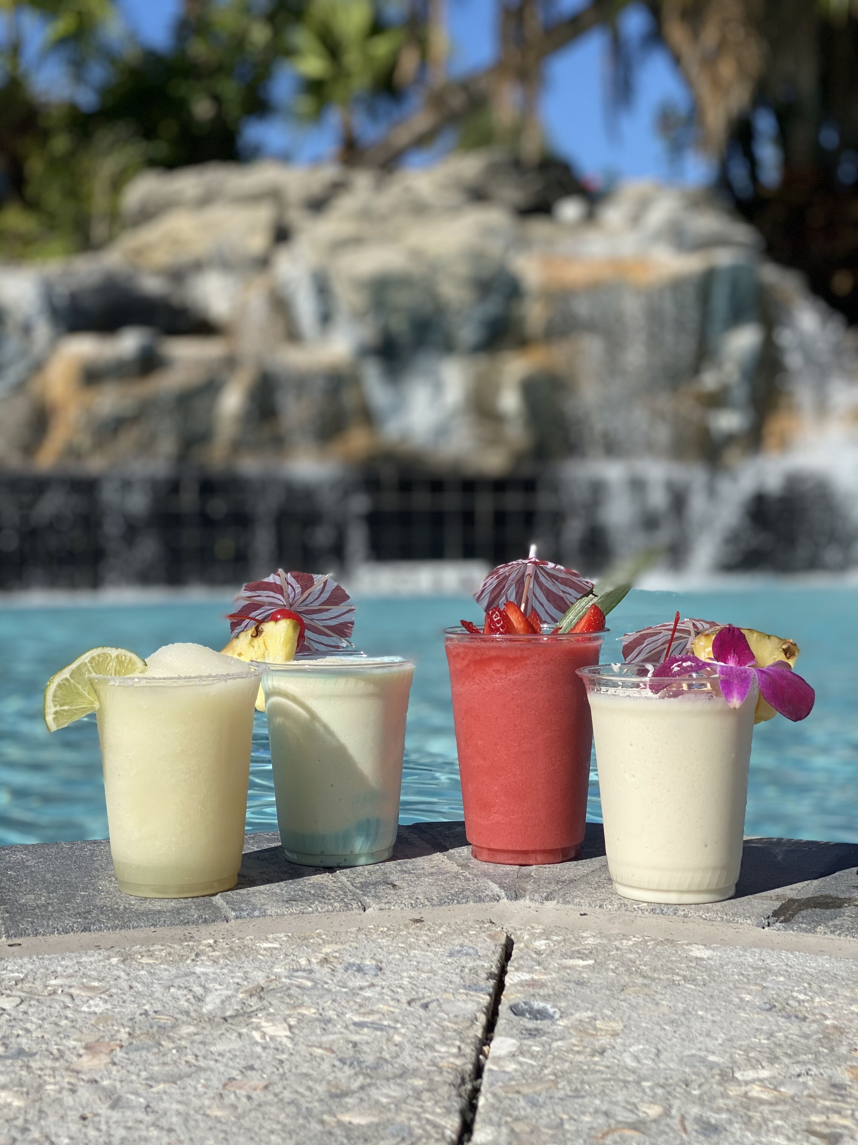 A poolside setting with four drinks in glasses, each garnished with fruits and a straw.