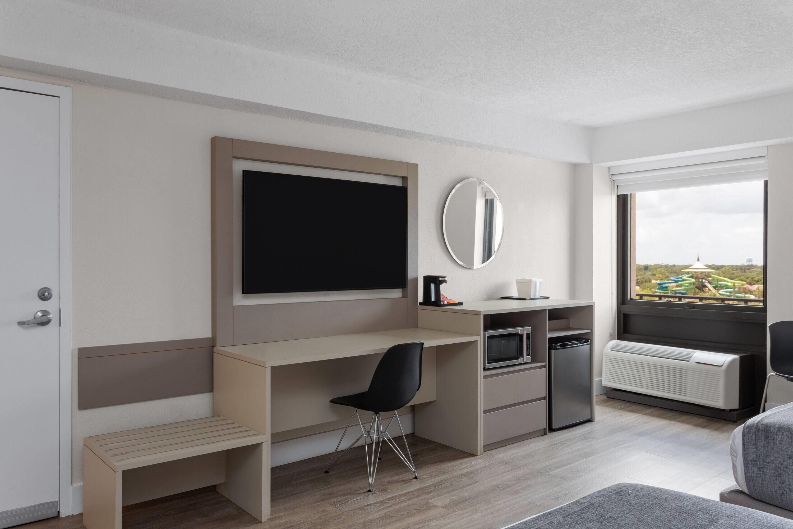 Een moderne hotelkamer met een bed, een bureau met stoel, een televisie, een spiegel, een kleine kitchenette en een raam met uitzicht.