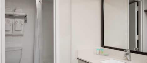 Une salle de bain comprenant des toilettes, une douche avec un rideau blanc, un lavabo avec un miroir et des articles de toilette posés sur le comptoir.