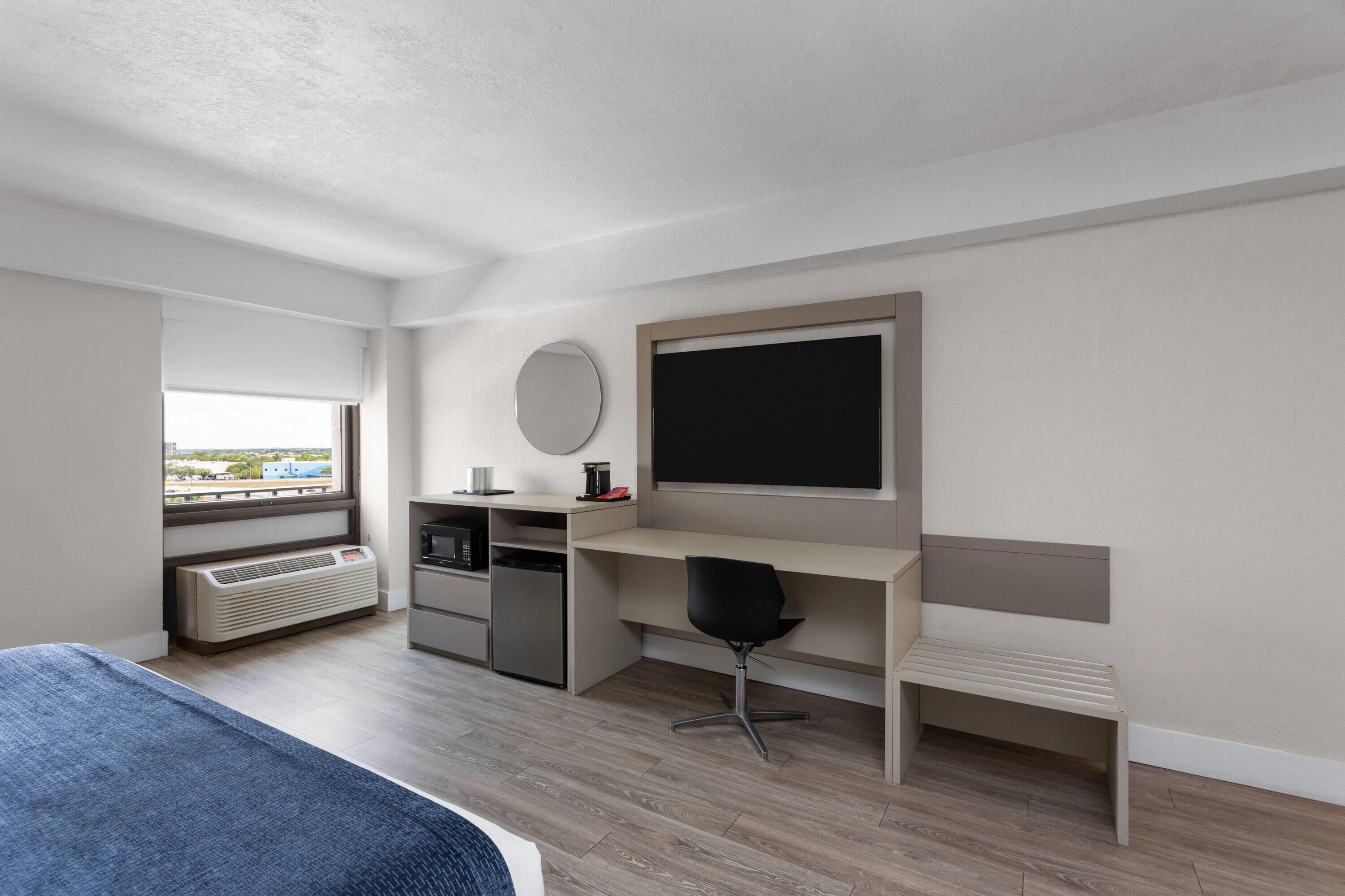 A modern hotel room with a bed, a desk, a chair, a heater, a window, and a view of the city.