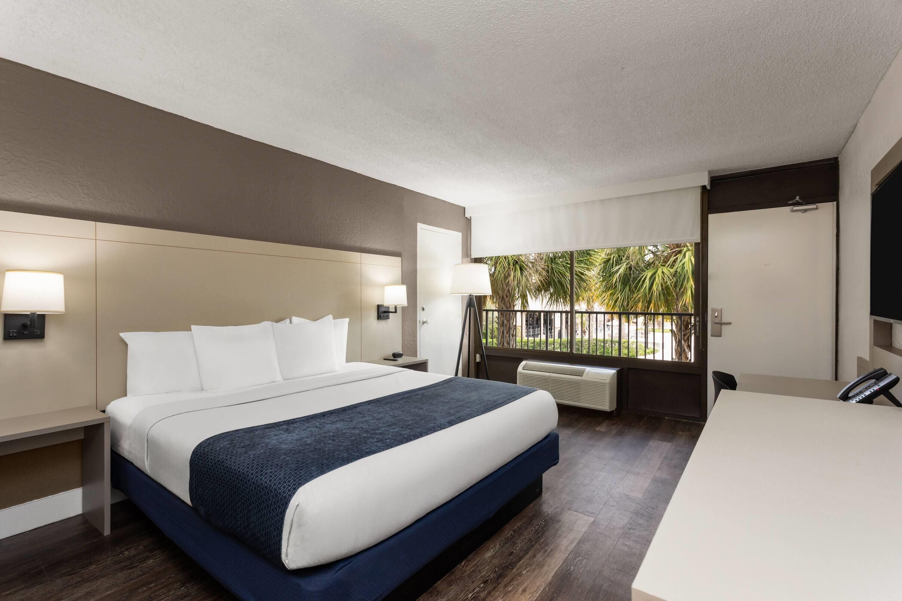 A hotel room with a large bed, bedside lamps, a desk, and a view of palm trees.