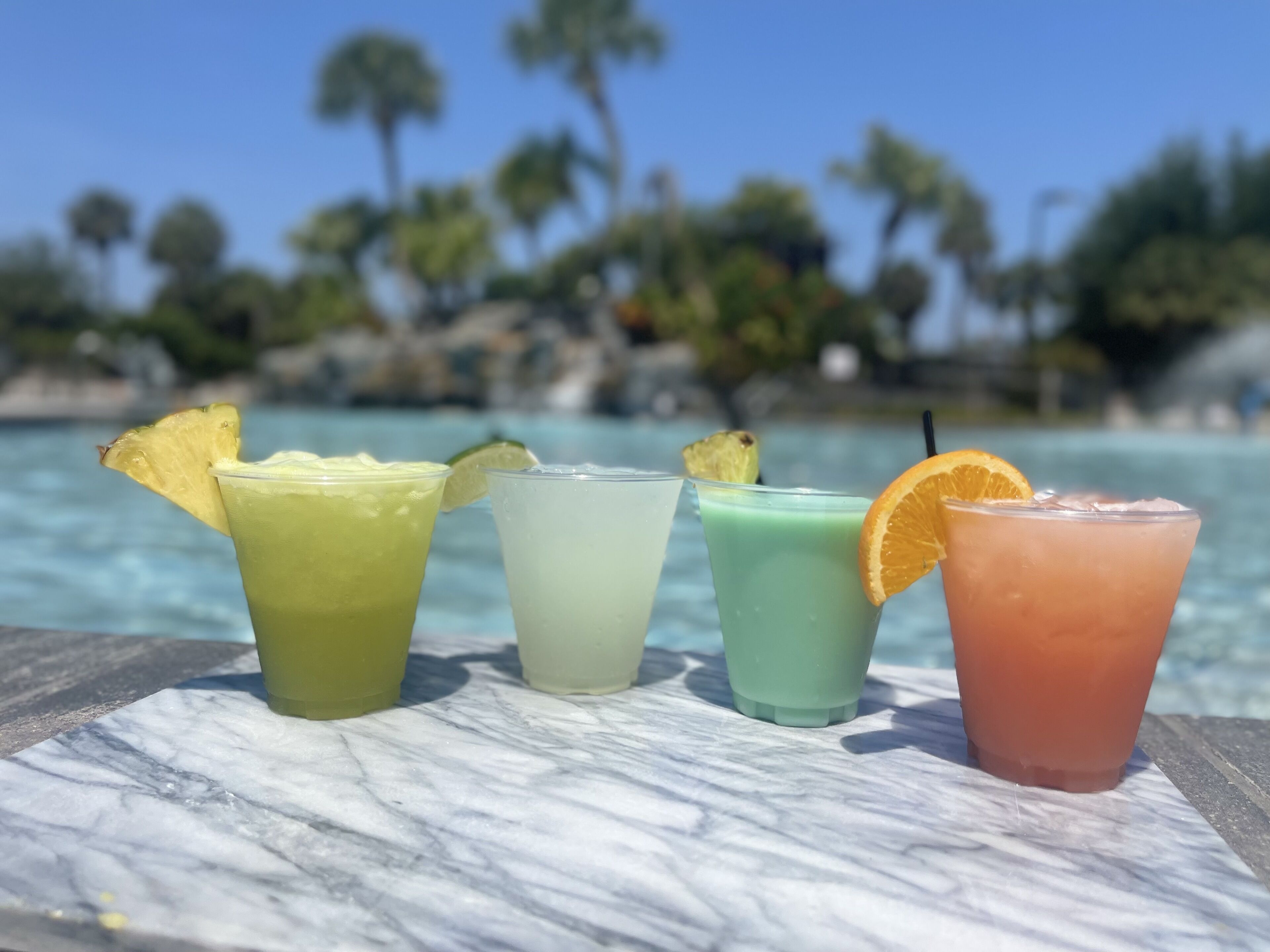 Four colorful drinks on a marble surface with a pool and palm trees in the background.