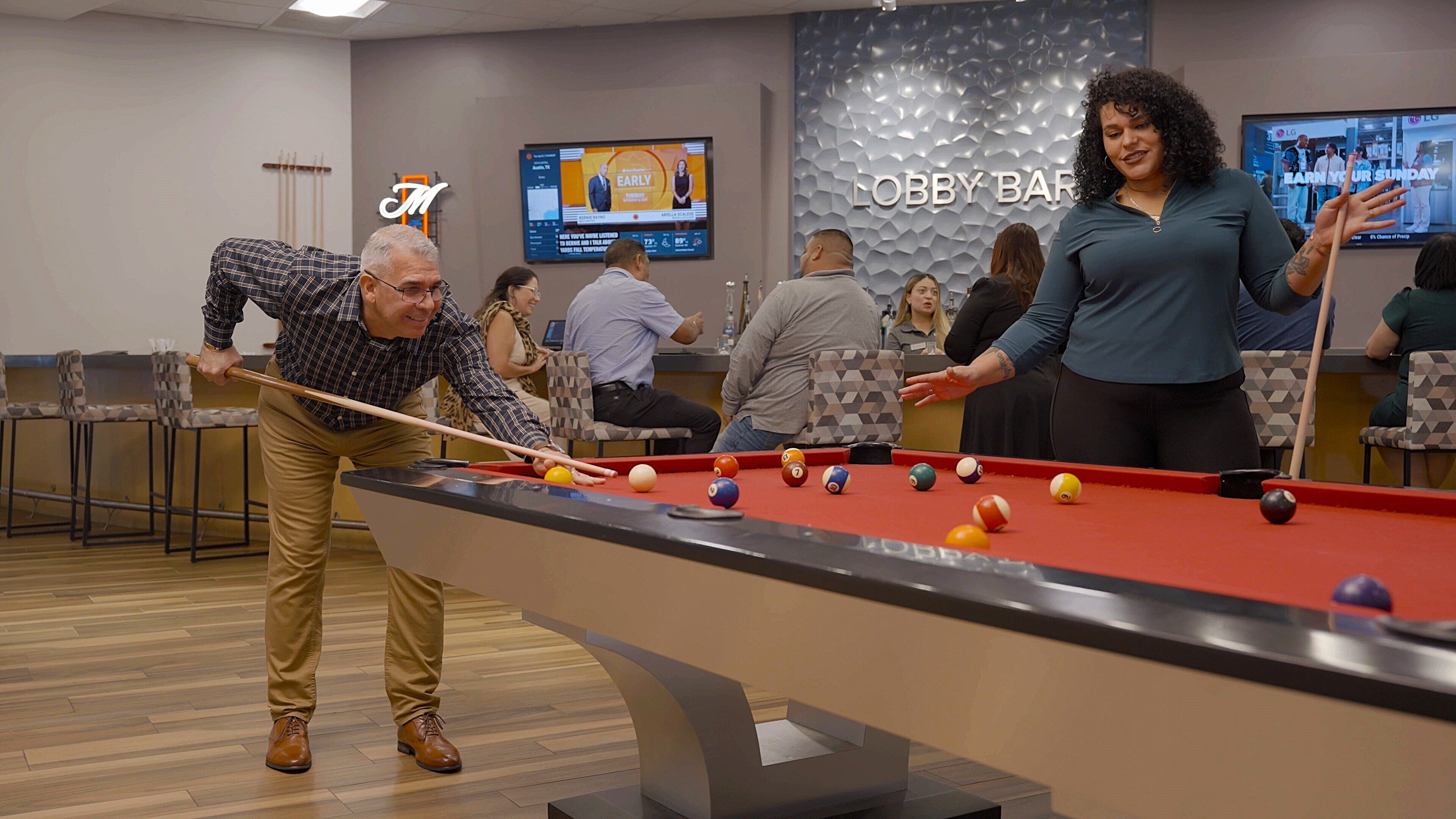 A pool table in a lobby area with a television displaying a news program and a sign reading "Lobby Bar".