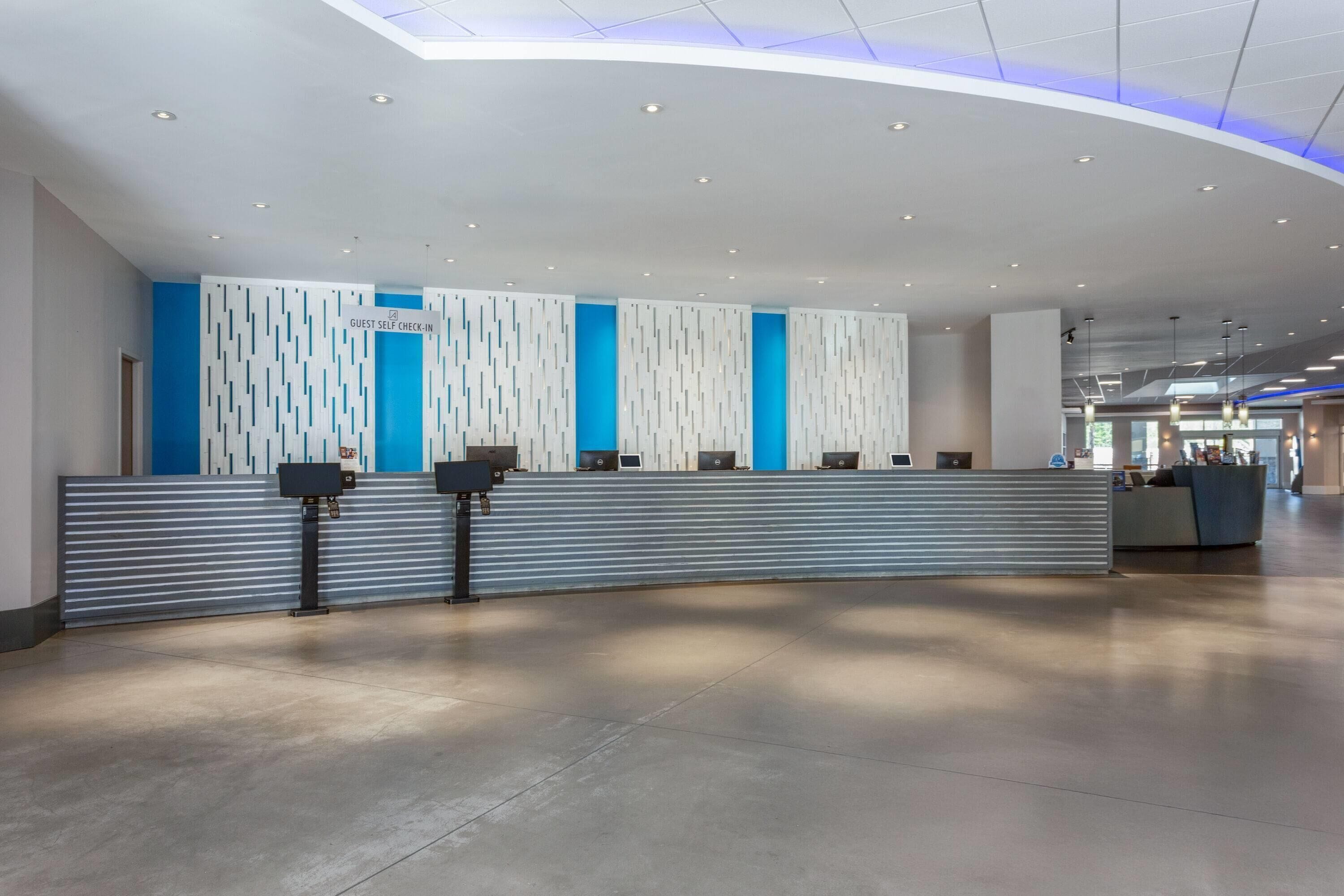 A modern hotel reception area with a curved reception desk, a 'Guest Self Check-In' sign, and a blue accent wall.